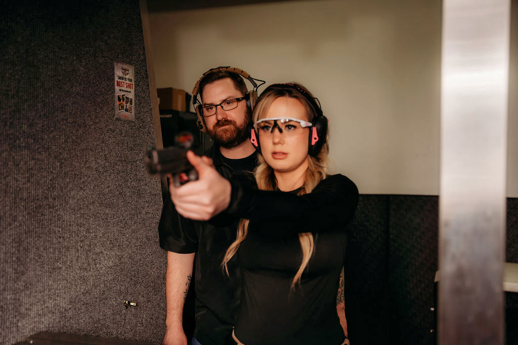 Woman shooting a pistol at an indoor range bay, with a man standing behind.